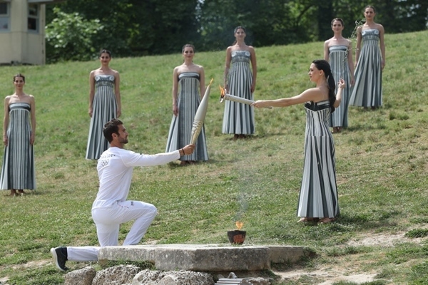 扮演最高女祭司的演員瑪麗·米娜（前右）點燃第一棒火炬手——希臘運動員、東京奧運會男子賽艇項目金牌得主斯特凡諾斯·恩圖斯科斯手中的火炬。新華社記者 趙丁喆 攝