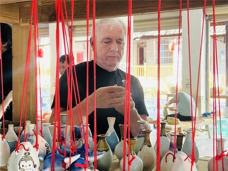 鄉(xiāng)愁如同一根根紅線(xiàn)，將游子與故鄉(xiāng)永遠(yuǎn)地牽連到一起?；逄账囄幕瘎?chuàng)意一條街的母氏陶藝館內(nèi)，紅線(xiàn)吊著陶瓶，思緒游走萬(wàn)里。摩爾多瓦“信息標(biāo)簽”通訊社社長(zhǎng)亞歷山大·塔納斯端詳著館內(nèi)展示的陶藝作品，并挑選出心儀的小擺件帶回摩爾多瓦，也帶走花茂村的“鄉(xiāng)愁”。（張亞欣攝）